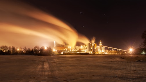 A nighttime industrial landscape with a factory emitting bright orange light and billowing smoke under a starry sky. The surrounding area is dark, and the contrast of light accentuates the structure's outlines against a backdrop of trees.