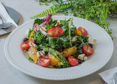 A plate filled with a colorful, fresh salad containing a variety of ingredients such as leafy greens, cherry tomatoes, orange slices, and cubes of cheese. The salad is garnished with a light dressing, and a set of utensils is placed on a napkin nearby. Lush greenery in the background enhances the freshness of the scene.