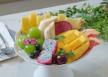 A beautifully arranged plate of sliced tropical fruits, including pineapple, banana, mango, grapefruit, and dragon fruit, is presented on a white pedestal dish. Green grapes, dark berries, and a cherry add variety. A garnish of fresh mint leaves and pink flowers enhances the display.