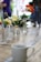 A wooden table adorned with various glass vases containing colorful flowers. A bottle of hand sanitizer is placed amongst the vases. In the foreground, there is a white ceramic mug, possibly containing a beverage. The background is blurred, with indistinct shapes and colors suggesting the presence of people and greenery.