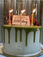 A birthday cake adorned with three lit candles and a 'Happy Birthday' plaque. The cake has a white base with green icing dripped from the top. Additional decorations include various colored frosting and fruits.