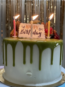 A birthday cake adorned with three lit candles and a 'Happy Birthday' plaque. The cake has a white base with green icing dripped from the top. Additional decorations include various colored frosting and fruits.