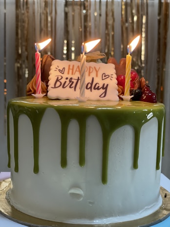 A birthday cake adorned with three lit candles and a 'Happy Birthday' plaque. The cake has a white base with green icing dripped from the top. Additional decorations include various colored frosting and fruits.