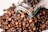 A clear glass jar is tipped over, spilling dark brown coffee beans onto a light surface. The coffee beans are of various sizes and shades of brown, appearing glossy and freshly roasted.