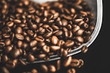 A collection of roasted coffee beans scattered against a dark background. The beans have a shiny, oily surface, indicating their freshness. They appear rich and textured, suggesting a robust aroma.