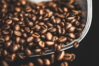 A collection of roasted coffee beans scattered against a dark background. The beans have a shiny, oily surface, indicating their freshness. They appear rich and textured, suggesting a robust aroma.