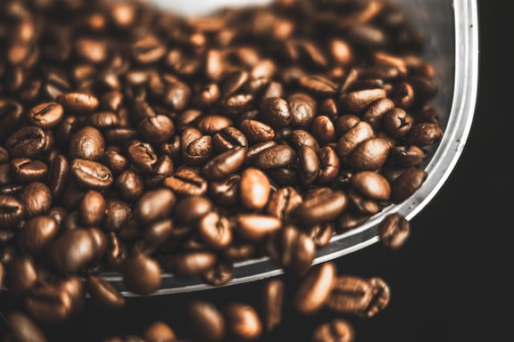 A collection of roasted coffee beans scattered against a dark background. The beans have a shiny, oily surface, indicating their freshness. They appear rich and textured, suggesting a robust aroma.