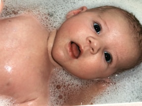 A baby with wide eyes and an open mouth is lying in a bath filled with foam and bubbles. The baby's skin glistens with water, and the foam surrounds its head.