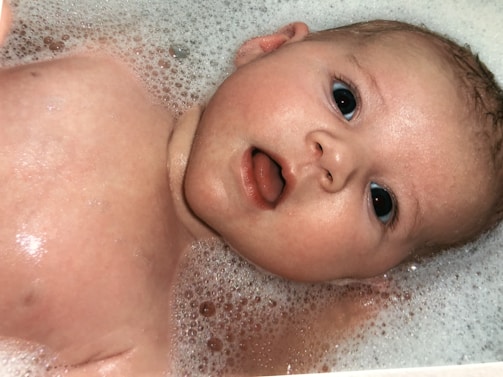A baby with wide eyes and an open mouth is lying in a bath filled with foam and bubbles. The baby's skin glistens with water, and the foam surrounds its head.