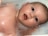A baby with wide eyes and an open mouth is lying in a bath filled with foam and bubbles. The baby's skin glistens with water, and the foam surrounds its head.