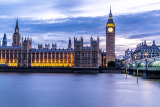 A majestic view of the Houses of Parliament and the iconic clock tower at dusk, with the River Thames in the foreground. The sky is filled with soft, blueish clouds and the building is illuminated with golden lights, reflecting in the water.