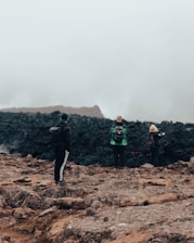 Three people are standing on rugged terrain, facing a dark, rocky area that is emitting smoke or steam. The atmosphere is misty and overcast, creating a sense of mystery and exploration.