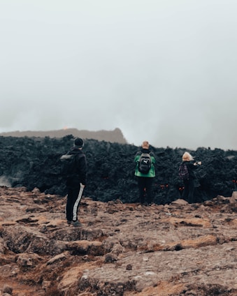 Three people are standing on rugged terrain, facing a dark, rocky area that is emitting smoke or steam. The atmosphere is misty and overcast, creating a sense of mystery and exploration.