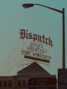 A large retro-style sign with ornate lettering reads 'Dispatch Ohio's Greatest Home Newspaper' along with a note of '152 Years of Service.' This sign is mounted on a metal framework above a historic building with architectural details visible. In the foreground, there is a street lamp and signs pointing toward Downtown, German Village, and a nearby interstate, creating a mix of historic and urban elements.