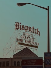 A large retro-style sign with ornate lettering reads 'Dispatch Ohio's Greatest Home Newspaper' along with a note of '152 Years of Service.' This sign is mounted on a metal framework above a historic building with architectural details visible. In the foreground, there is a street lamp and signs pointing toward Downtown, German Village, and a nearby interstate, creating a mix of historic and urban elements.