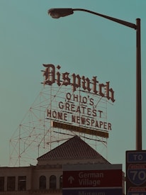 A large retro-style sign with ornate lettering reads 'Dispatch Ohio's Greatest Home Newspaper' along with a note of '152 Years of Service.' This sign is mounted on a metal framework above a historic building with architectural details visible. In the foreground, there is a street lamp and signs pointing toward Downtown, German Village, and a nearby interstate, creating a mix of historic and urban elements.