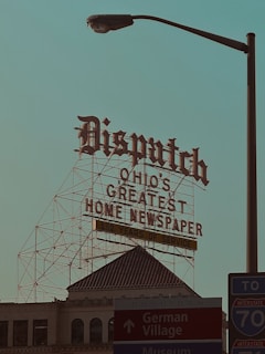 A large retro-style sign with ornate lettering reads 'Dispatch Ohio's Greatest Home Newspaper' along with a note of '152 Years of Service.' This sign is mounted on a metal framework above a historic building with architectural details visible. In the foreground, there is a street lamp and signs pointing toward Downtown, German Village, and a nearby interstate, creating a mix of historic and urban elements.