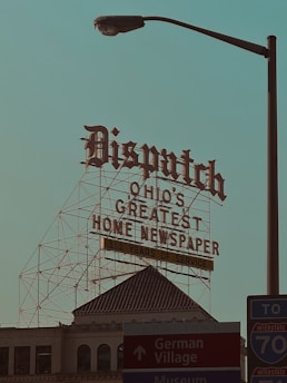 A large retro-style sign with ornate lettering reads 'Dispatch Ohio's Greatest Home Newspaper' along with a note of '152 Years of Service.' This sign is mounted on a metal framework above a historic building with architectural details visible. In the foreground, there is a street lamp and signs pointing toward Downtown, German Village, and a nearby interstate, creating a mix of historic and urban elements.