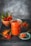 A glass jar filled with red chili sauce is placed on a dark slate surface, accompanied by a small bowl containing whole red chilies. A spoon rests on the jar's lid, with a portion of the sauce on it. Chili powder is sprinkled on the surface, adding texture and color.