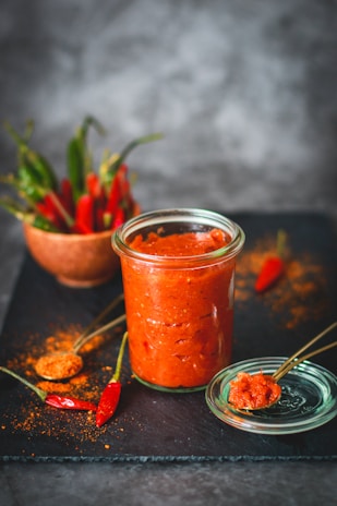 A glass jar filled with red chili sauce is placed on a dark slate surface, accompanied by a small bowl containing whole red chilies. A spoon rests on the jar's lid, with a portion of the sauce on it. Chili powder is sprinkled on the surface, adding texture and color.