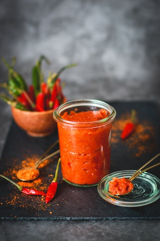 A glass jar filled with red chili sauce is placed on a dark slate surface, accompanied by a small bowl containing whole red chilies. A spoon rests on the jar's lid, with a portion of the sauce on it. Chili powder is sprinkled on the surface, adding texture and color.