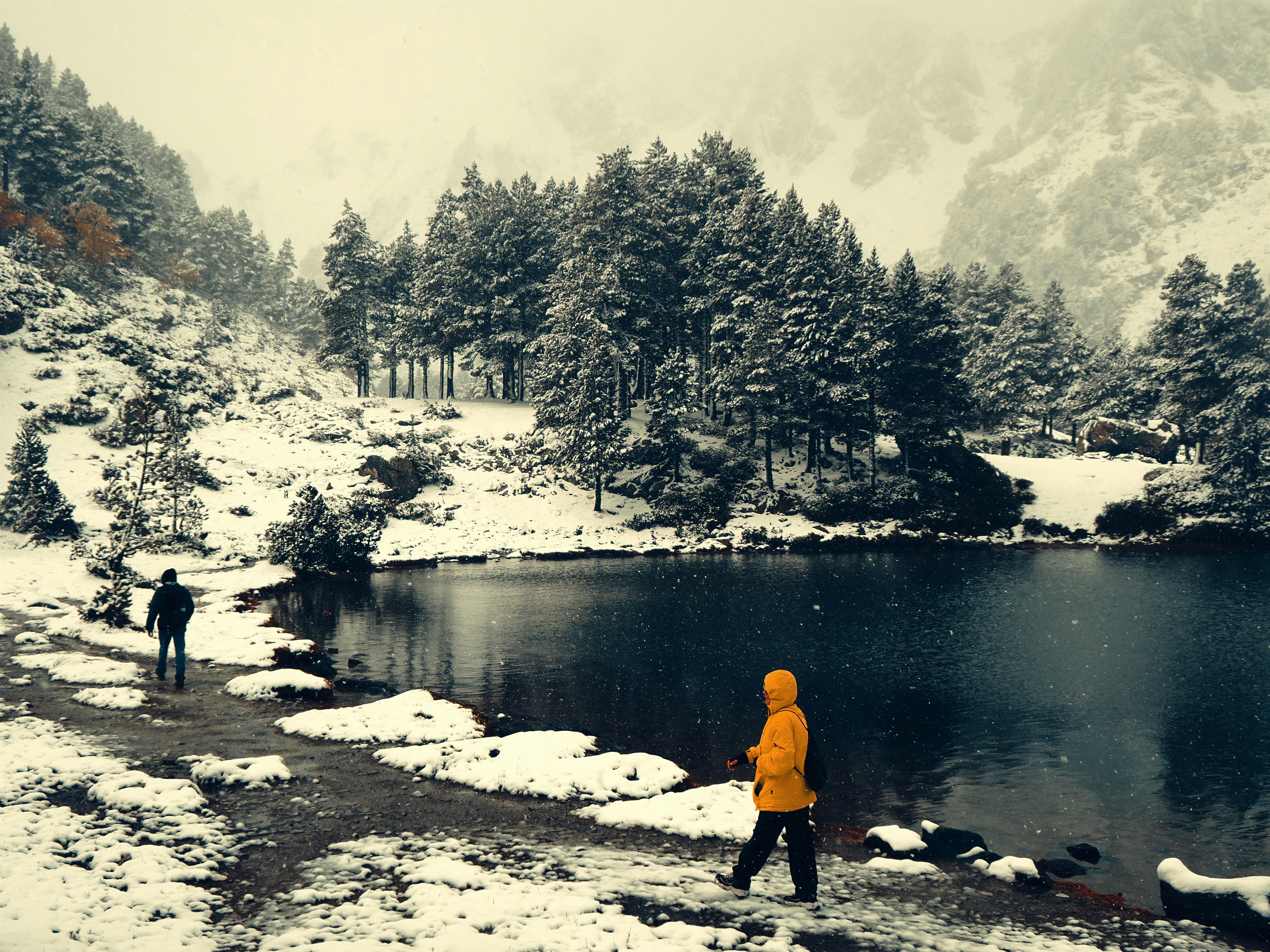 Snow-dusted lakeside landscape photograph with a bright yellow-jacketed figure at the water's edge, framed by pine trees and a second hiker along the shore.