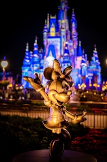 A golden statue of a cheerful cartoon mouse character stands in front of a beautifully illuminated castle. The castle is adorned in vibrant blue and purple lights, creating a magical and festive atmosphere. Lush gardens and glowing lamps accentuate the night scene.