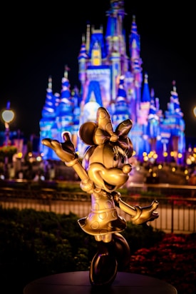 A golden statue of a cheerful cartoon mouse character stands in front of a beautifully illuminated castle. The castle is adorned in vibrant blue and purple lights, creating a magical and festive atmosphere. Lush gardens and glowing lamps accentuate the night scene.