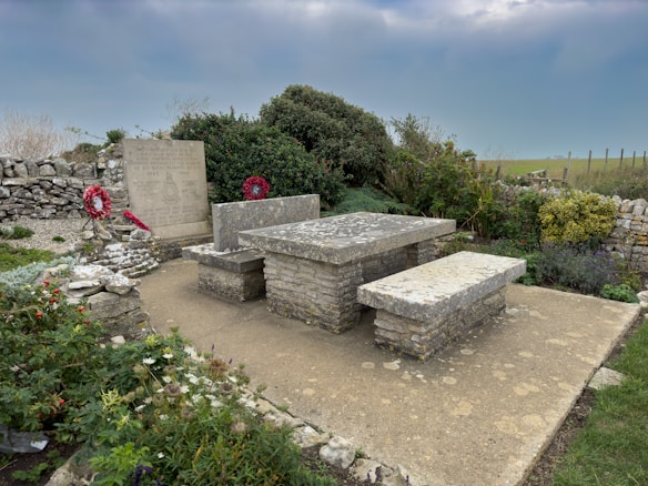 The image captures a stone memorial surrounded by a low stone wall. A large rectangular stone slab and two stone benches suggest a memorial site. Two red wreaths are placed against the inscribed memorial plaque, and there are green bushes and flowering plants nearby. The setting appears serene, with a cloudy sky and grassy landscape in the background.