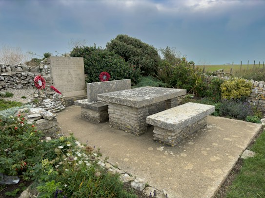 The image captures a stone memorial surrounded by a low stone wall. A large rectangular stone slab and two stone benches suggest a memorial site. Two red wreaths are placed against the inscribed memorial plaque, and there are green bushes and flowering plants nearby. The setting appears serene, with a cloudy sky and grassy landscape in the background.