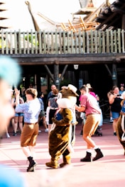 A group of people, some in costumes, are engaging in a lively dance routine in an outdoor setting. The foreground features a character in a brown animal costume with a large head, surrounded by performers in casual and colorful attire. The scene captures a festive atmosphere with a wooden fence and structures in the background.