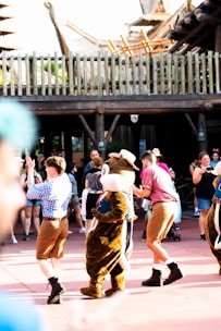 A group of people, some in costumes, are engaging in a lively dance routine in an outdoor setting. The foreground features a character in a brown animal costume with a large head, surrounded by performers in casual and colorful attire. The scene captures a festive atmosphere with a wooden fence and structures in the background.