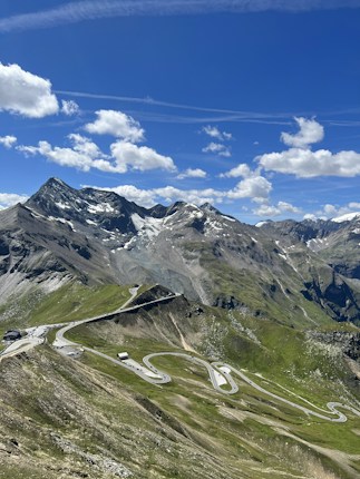 A winding road snakes through a lush green mountainous landscape with rugged peaks in the background. Snow-capped mountains are visible under a clear blue sky scattered with a few fluffy white clouds.