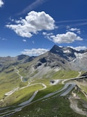 A picturesque mountain landscape featuring a winding road cutting through lush green hills. The scene is dominated by a towering, snow-capped peak under a vivid blue sky filled with fluffy white clouds. People can be seen walking along paths, enjoying the scenic view.