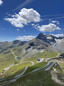 A picturesque mountain landscape featuring a winding road cutting through lush green hills. The scene is dominated by a towering, snow-capped peak under a vivid blue sky filled with fluffy white clouds. People can be seen walking along paths, enjoying the scenic view.