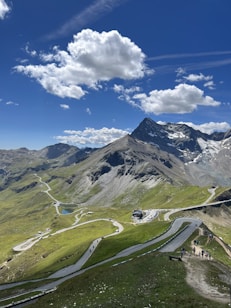 A picturesque mountain landscape featuring a winding road cutting through lush green hills. The scene is dominated by a towering, snow-capped peak under a vivid blue sky filled with fluffy white clouds. People can be seen walking along paths, enjoying the scenic view.