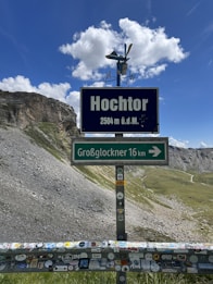 A mountain pass with a sign reading 'Hochtor 2504 m ü.d.M.' indicating elevation and a directional sign pointing towards Großglockner, located in a rocky, mountainous landscape with a clear blue sky and scattered clouds. The foreground includes a barrier covered in various stickers.