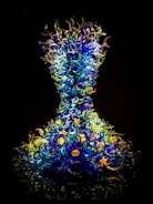 A vibrant and intricate glass sculpture composed of numerous swirling, curly elements in shades of blue, yellow, and green. The structure resembles an hourglass form, with the glass components appearing almost luminous against a dark background.
