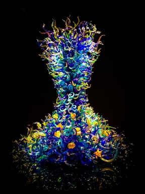 A vibrant and intricate glass sculpture composed of numerous swirling, curly elements in shades of blue, yellow, and green. The structure resembles an hourglass form, with the glass components appearing almost luminous against a dark background.