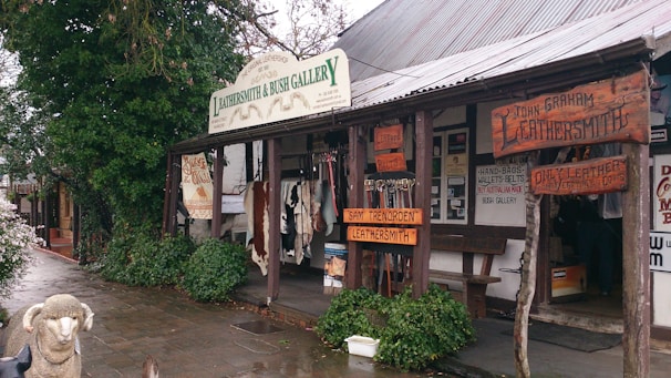 A rustic storefront showcasing handcrafted leather goods, with a wooden overhang displaying various signs for a leathersmith gallery. The exterior features animal hides and leather belts on display. The area is surrounded by greenery and has a wet cobblestone pavement. A decorative sheep statue is positioned in front of the store.