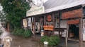 A rustic storefront showcasing handcrafted leather goods, with a wooden overhang displaying various signs for a leathersmith gallery. The exterior features animal hides and leather belts on display. The area is surrounded by greenery and has a wet cobblestone pavement. A decorative sheep statue is positioned in front of the store.