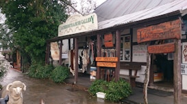 A rustic storefront showcasing handcrafted leather goods, with a wooden overhang displaying various signs for a leathersmith gallery. The exterior features animal hides and leather belts on display. The area is surrounded by greenery and has a wet cobblestone pavement. A decorative sheep statue is positioned in front of the store.