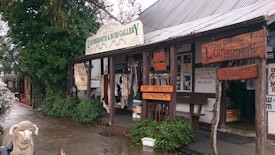 A rustic storefront showcasing handcrafted leather goods, with a wooden overhang displaying various signs for a leathersmith gallery. The exterior features animal hides and leather belts on display. The area is surrounded by greenery and has a wet cobblestone pavement. A decorative sheep statue is positioned in front of the store.