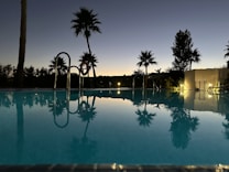 A serene swimming pool at dusk, with silhouettes of palm trees reflecting on the calm water. The sky is transitioning from day to night with a gradient of blues and oranges. Dim lights illuminate the surroundings, enhancing the tranquil atmosphere.