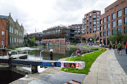 A serene canal scene in an urban area featuring brick buildings and people casually strolling or sitting by the water. The canal is bordered by a decoratively painted lock, and there is a mix of historic and modern architecture. People are enjoying the outdoor space, reflecting a lively social atmosphere.