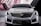 A sleek and modern car with a prominent front grille, angular headlights, and a smooth hood design. The car is set against a sophisticated backdrop that features geometric shapes and lines.
