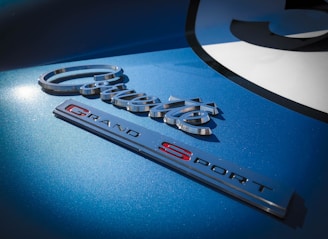 A close-up view of car branding emblems on a blue metallic surface featuring the words 'Corvette' and 'Grand Sport' with a decorative font. The lighting highlights the sleek design and reflective finish of the emblems.