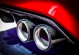 A close-up of a car's dual exhaust pipes with a carbon fiber finish, highlighted against a glossy red body. The setting emphasizes the intricate details and the metallic sheen of the exhaust tips.