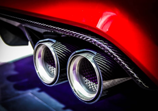 A close-up of a car's dual exhaust pipes with a carbon fiber finish, highlighted against a glossy red body. The setting emphasizes the intricate details and the metallic sheen of the exhaust tips.
