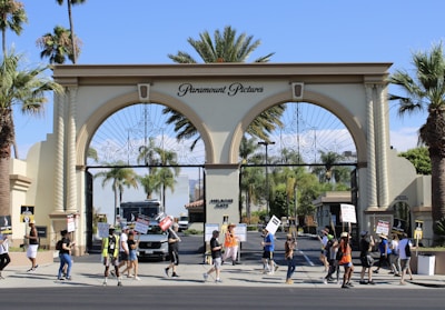 A group of people are holding signs and picketing in front of a large ornate gate with three arches labeled Paramount Pictures. The scene includes palm trees, an entrance sign reading Melrose Gate, and a security vehicle parked inside the gate. The weather appears clear and sunny.